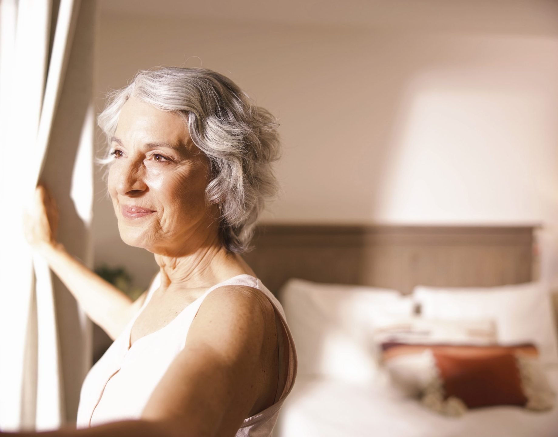An older woman with short gray hair stands by a window, gently holding open white curtains. Sunlight shines on her face, and there is a neatly made bed in the softly lit background.