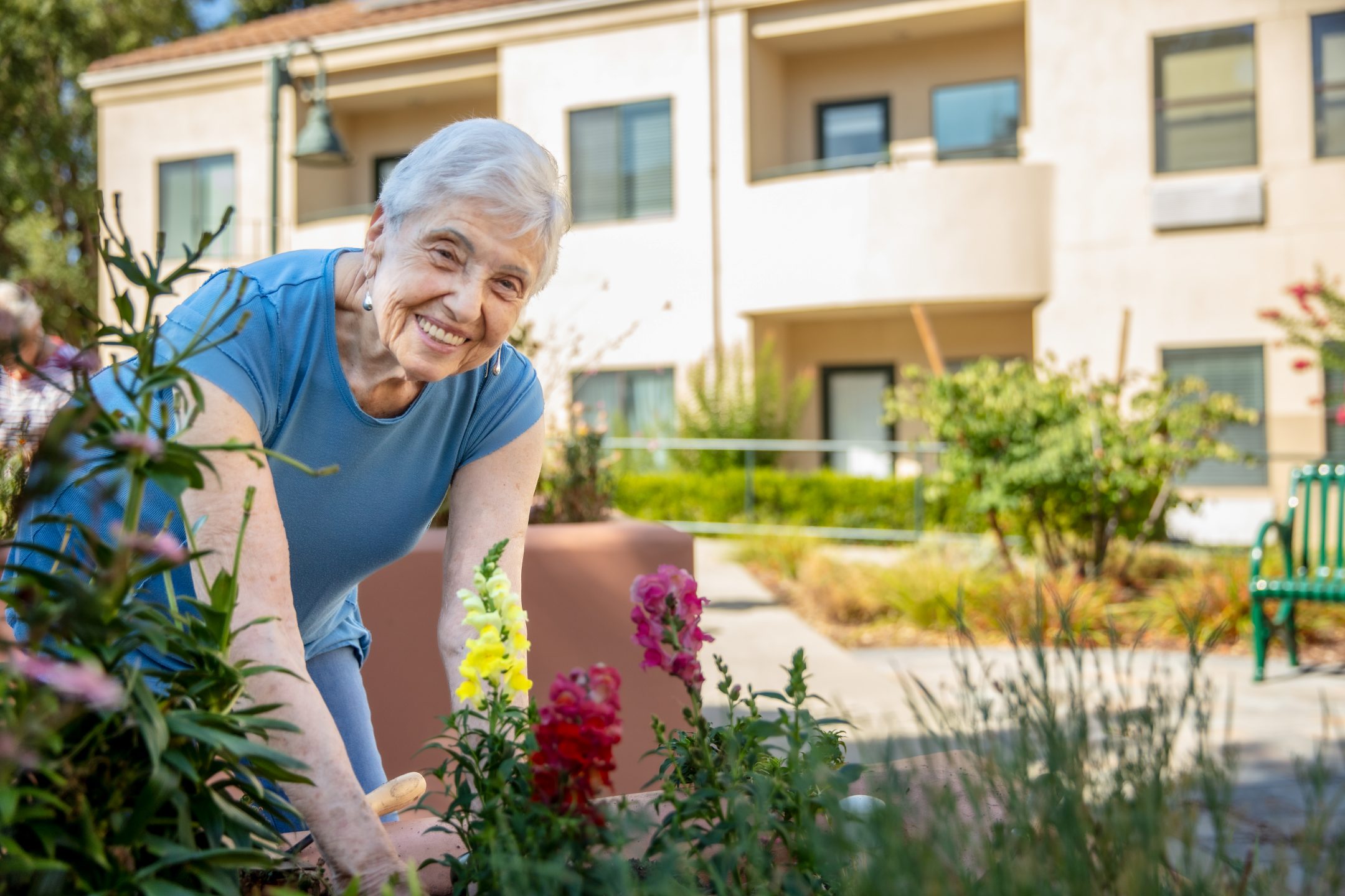 An elderly woman with short gray hair, wearing a blue shirt, smiles while tending to colorful flowers in a garden outside a residential building on a sunny day.