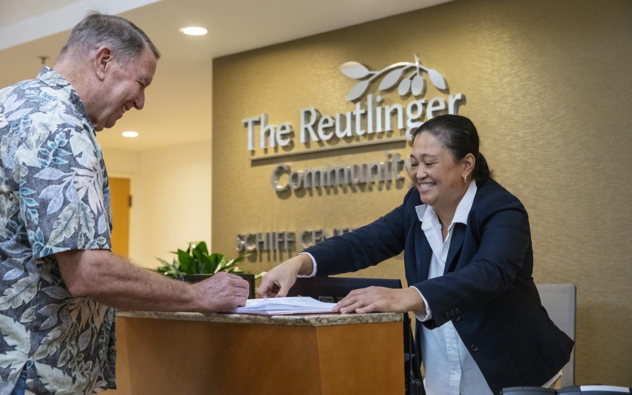 A man in a floral shirt signs papers at a reception desk while a smiling woman in a blazer assists him. The background displays a sign reading The Reutlinger Community. The Reutlinger Amenities and Services