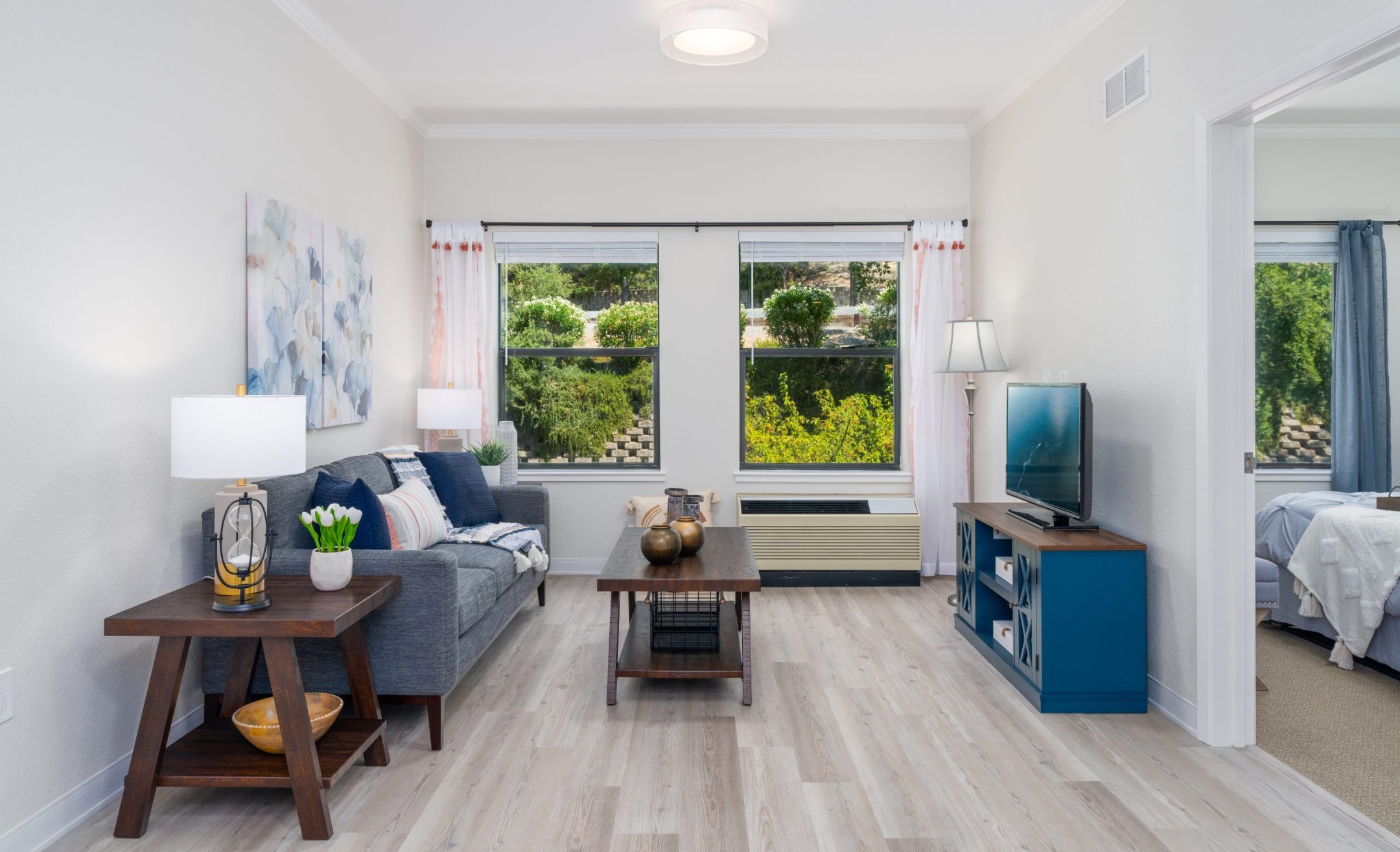 A bright living room with a gray sofa, wooden tables, and decor features two large windows with white curtains—showcasing the inviting comfort found in The Reutlinger living options. Light wood floors and a blue TV stand complete the look.
