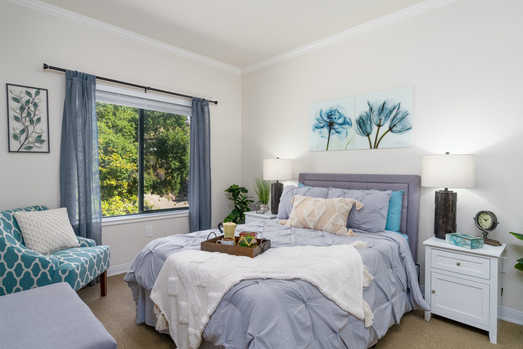 A cozy bedroom with a neatly made bed in gray and white bedding, two nightstands with lamps, a blue patterned chair, and blue floral artwork above the bed—one of the inviting spaces among The Reutlinger living options.