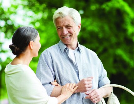 An older couple stands outdoors in a lush green park, smiling warmly at each other. The man holds a white picnic basket with bread inside, while the woman gently touches his arm.