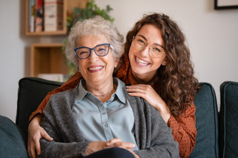 An older woman with gray hair and glasses sits on a couch, smiling, while a younger woman with curly hair and glasses hugs her from behind. Both appear happy and are looking at the camera.