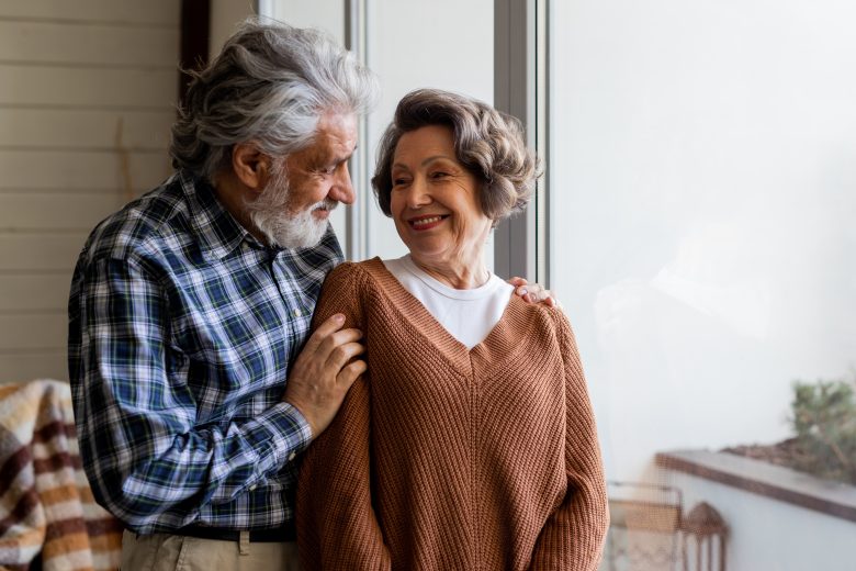 An elderly couple stands by a large window, smiling warmly at each other in their cozy, sunlit room—reflecting the happiness and contentment offered by The Reutlinger living options.