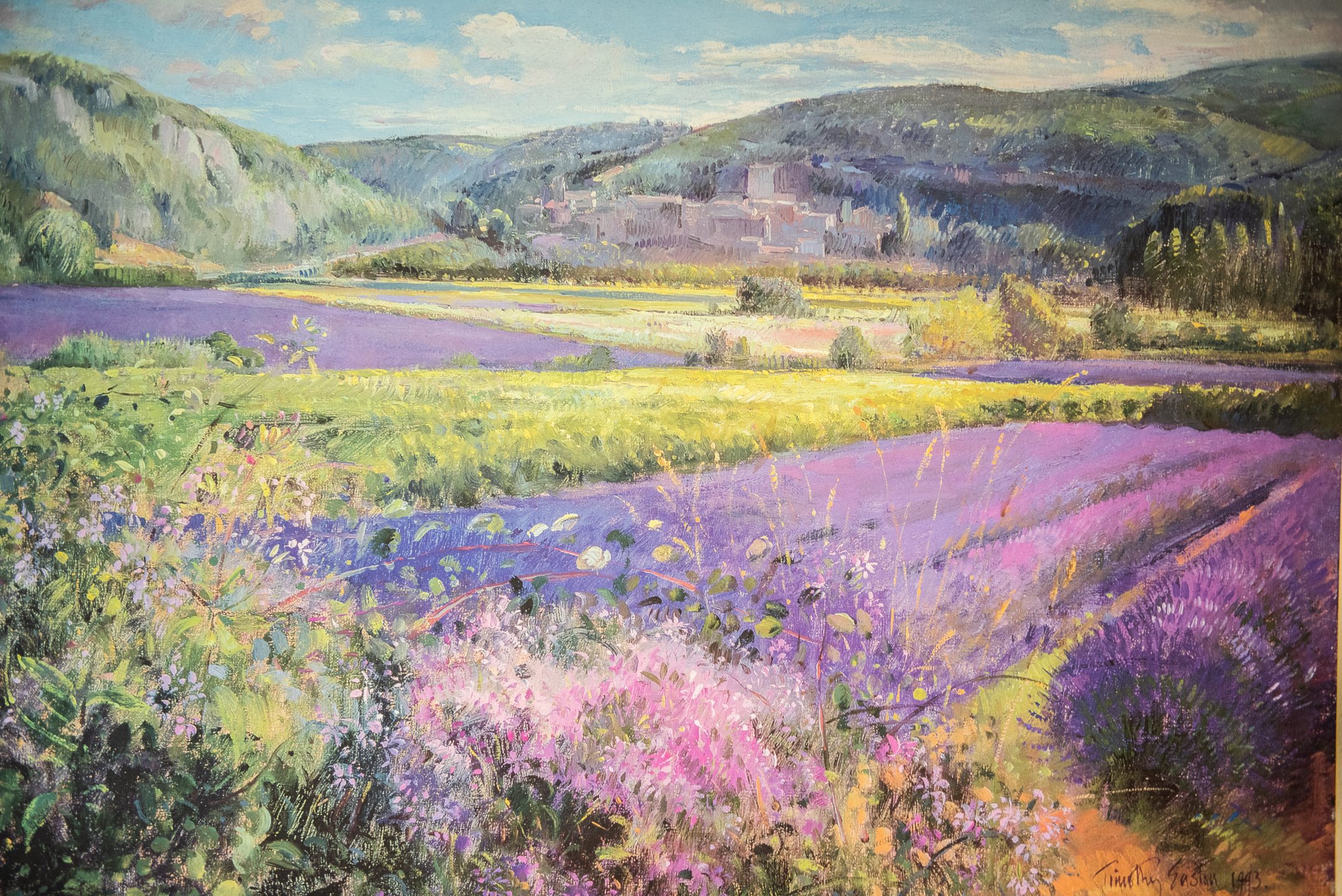 A vibrant painting of a countryside landscape captures the lifestyle of rural serenity with blooming lavender fields, green hills, and a distant village under a bright blue sky. The foreground bursts with colorful wildflowers.